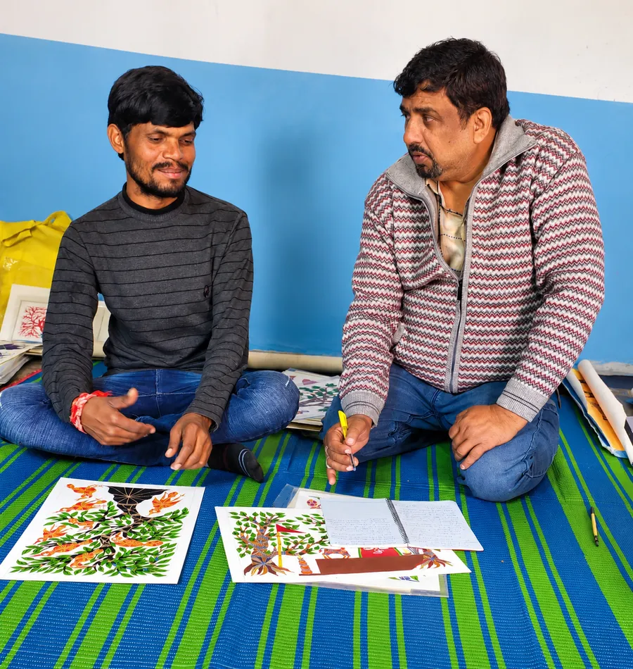 Two men sit on a striped mat, reviewing folk paintings laid out on the floor between them.