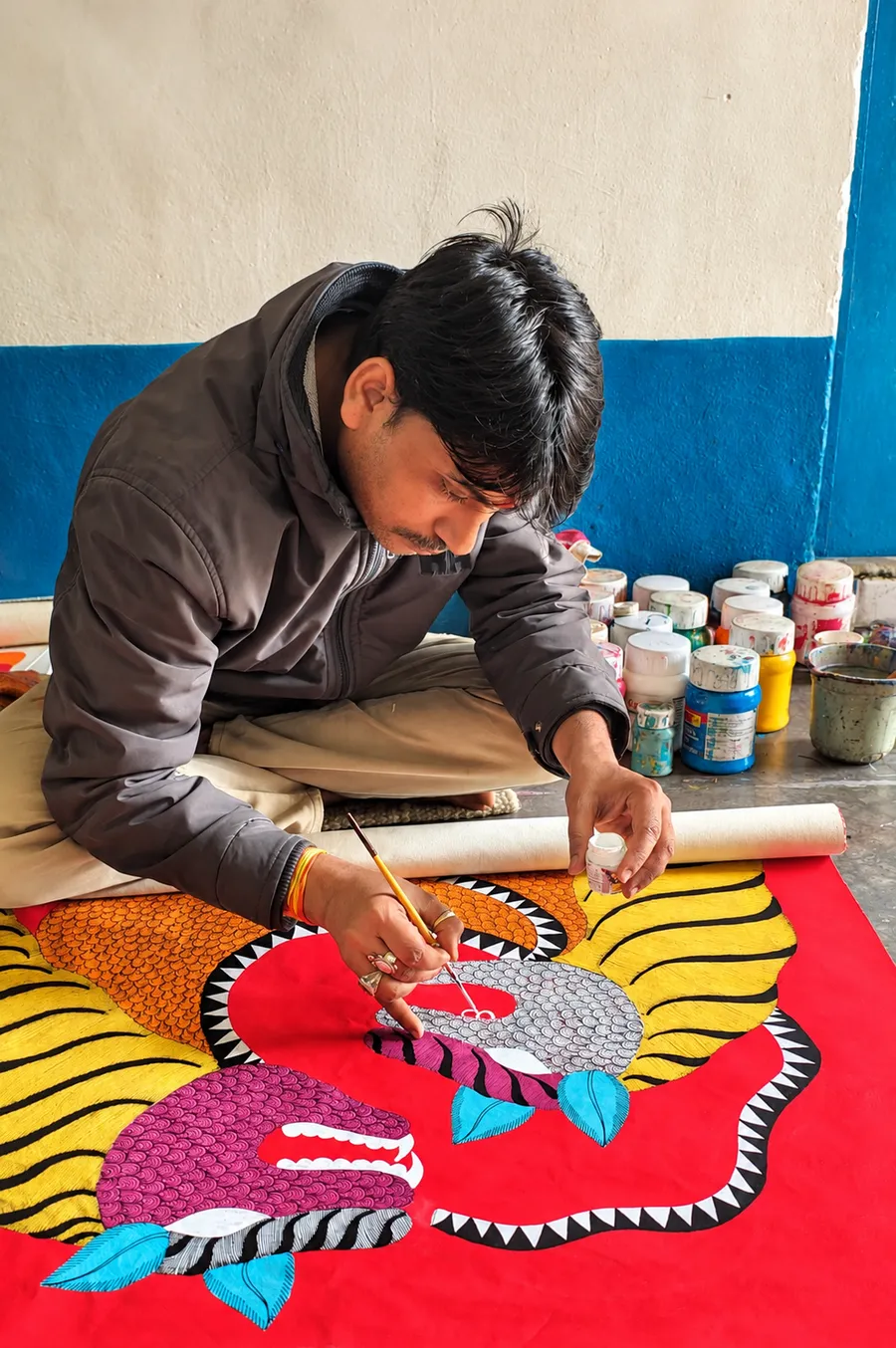 A young artist kneels on the floor painting a large, brightly coloured folk artwork, with paint jars beside him.