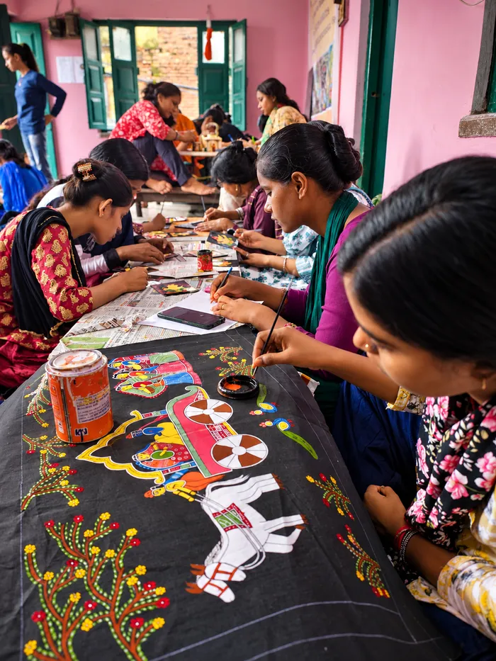Young women artists paint figures and trees on a long black cloth at a shared workshop table.
