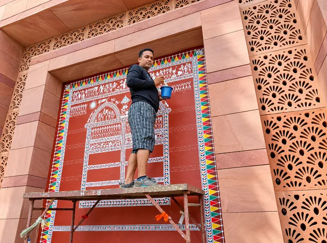 Rabinath Sabar stands on a scaffold beside a large Saura mural on a red wall.
