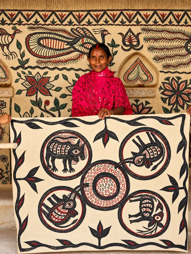 Rudhan Devi smiles as she holds up a large painting of animals in circles, in front of a wall painted with peacocks and flowers.
