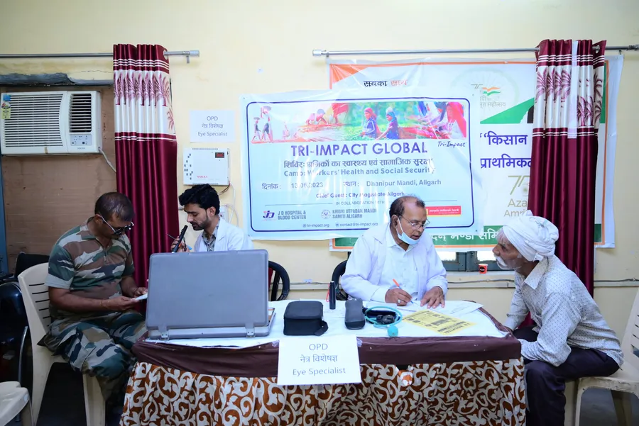 An eye specialist and camp staff sit at a table with workers under a Tri-Impact Global banner.